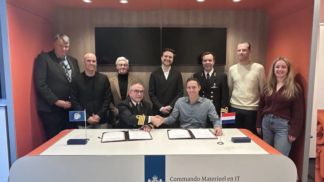  2 heren achter een desk, van wie 1 in militair DT,  schudden elkaar de hand met daarachter 6 mannen en een vrouw.