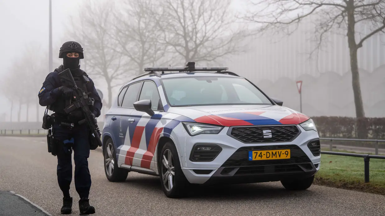 Een zwaarbewapende marechaussee in donker uniform staat naast een witte politieauto met rode-blauwe strepen en het kenteken "74-DMV-9" op een mistige weg.