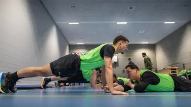 Jongeren in gele hesjes voeren diverse sportonderdelen uit in een sportzaal. 