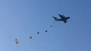 Tegen een strakblauwe lucht vliegt een donker militair transportvliegtuig. Erachter dalen meerdere parachutes met lading in een rij af.