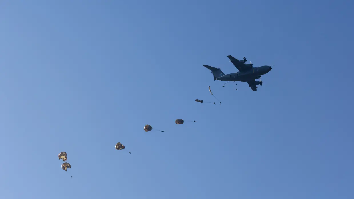 Tegen een strakblauwe lucht vliegt een donker militair transportvliegtuig. Erachter dalen meerdere parachutes met lading in een rij af.