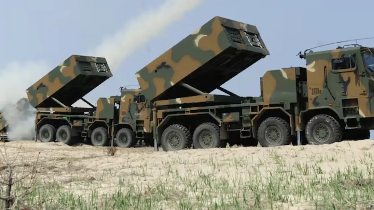 Een groot militair raketwerpervoertuig met camouflagekleuren schiet raketten af, met rook en vuur zichtbaar, op een zanderige heuvel met groen gras. 