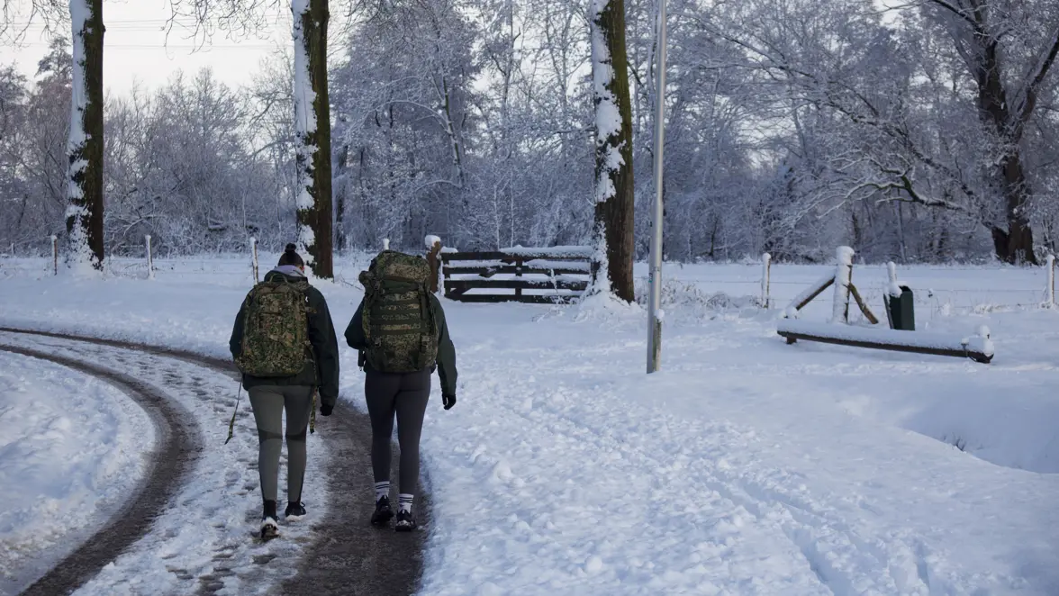 Twee mensen lopen met rugzakken in militaire print over een besneeuwd, rustig pad in een winterbos, omringd door sneeuwbedekte bomen en een bankje. 