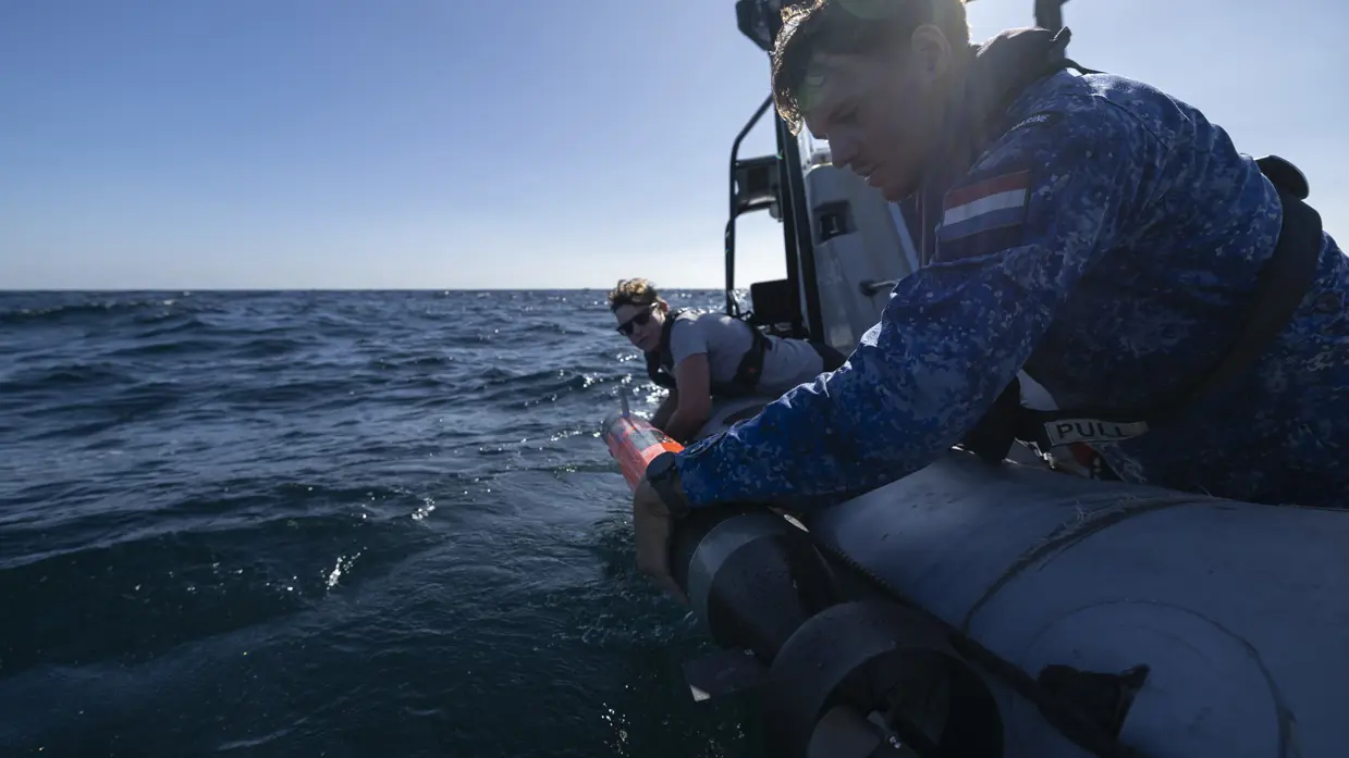 Een vrouw in grijs t-shirt en man in blauw uniform laten een oranjekleurige torpedo te water vanaf de RHIB-motorboot. 