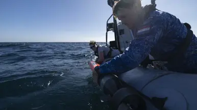 Een vrouw in grijs t-shirt en man in blauw uniform laten een oranjekleurige torpedo te water vanaf de RHIB-motorboot. 