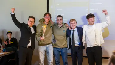 Vijf mannen van wie twee in net pak poseren met een bokaal voor een groot scherm.