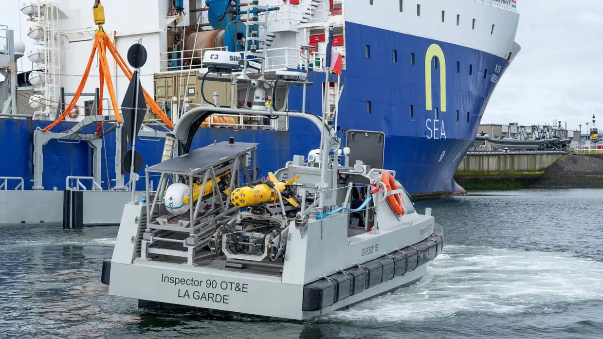 Links een grijs bootje met achterop verschillende drones. Dit vaart langs een groot vrachtschip. Daarnaast een gele, raketvormige drone met voorop een witte bol. Deze is ook te zien achterop het varende bootje.