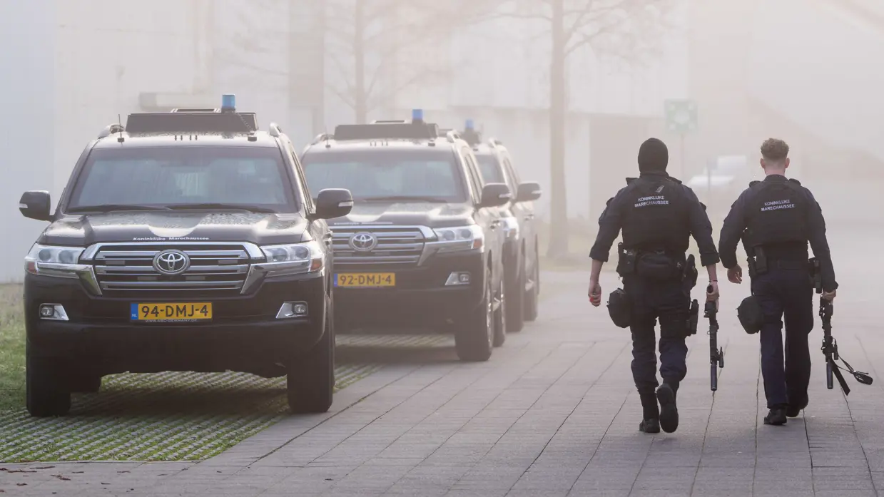 Twee marechaussees lopen op een betegeld pad. Ze dragen donkerblauwe uniformen met "Koninklijke Marechaussee" op de rug en zijn bewapend. Achter hen rijden drie zwarte, zware terreinwagens van de Koninklijke Marechaussee