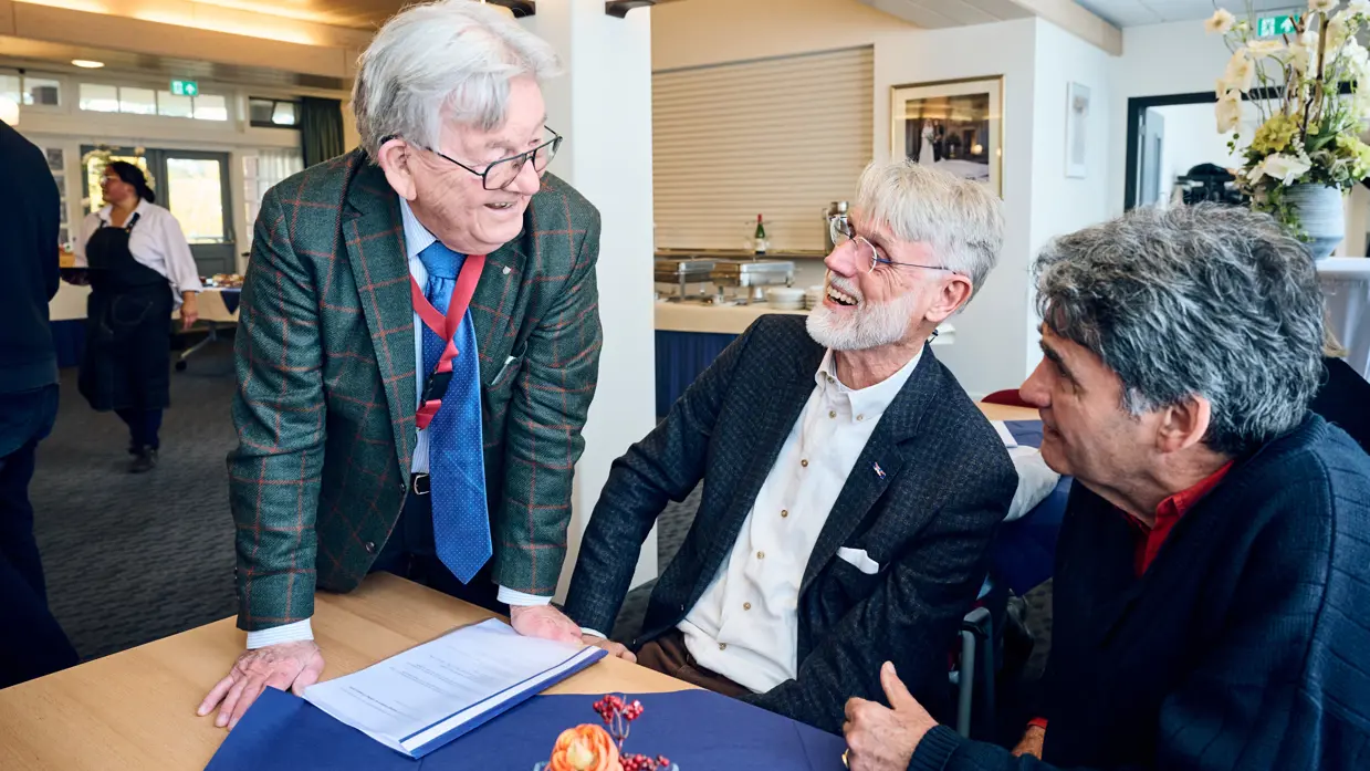 Een man in pak spreekt met twee heren die aan een tafel zitten in een zaal.