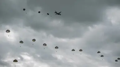 Onder een grijze bewolkte lucht dalen parachutisten met groene parachutes af. Op de achtergrond vliegt het vliegtuig waaruit ze sprongen.