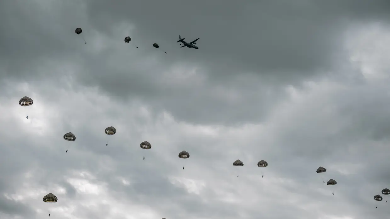 Onder een grijze bewolkte lucht dalen parachutisten met groene parachutes af. Op de achtergrond vliegt het vliegtuig waaruit ze sprongen.