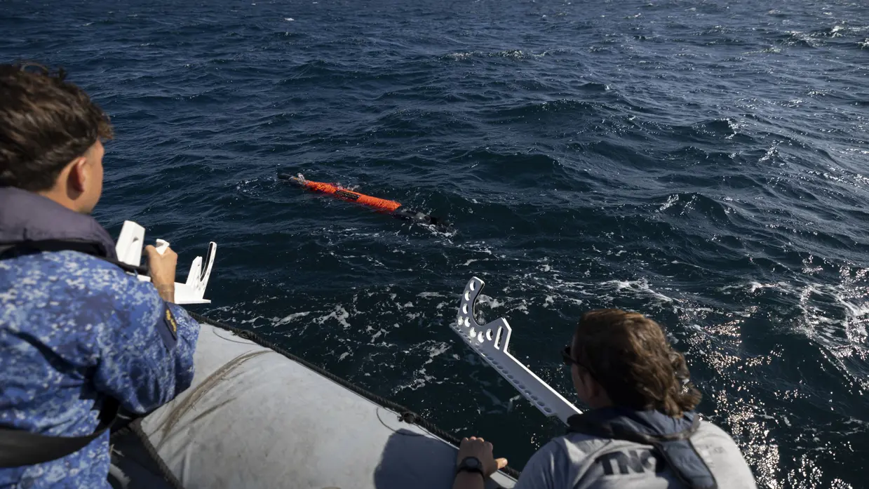 Twee personen kijken vanuit een motorbootje toe hoe een oranjekleurige torpedo in het water verdwijnt.