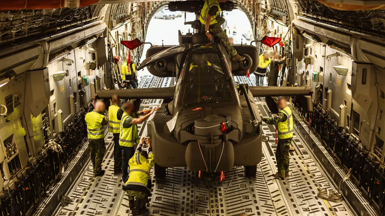 Mensen in gele hesje staan bij een Apache die nog in het laadruim van het transportvliegtuig staat.