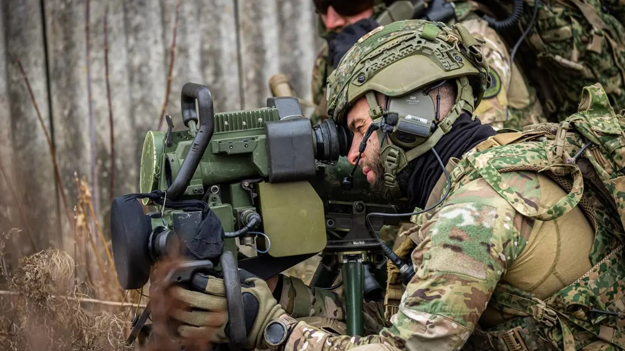 Een militair ligt achter een antitankwapen en kijkt met één oog door het richtmiddel. Achter hem is een collega te zien.
