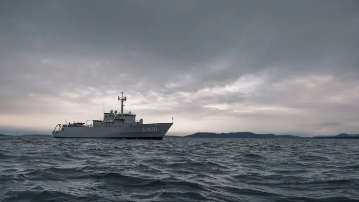 Een grijs schip vaart op zee met een kustlijn in de verte 