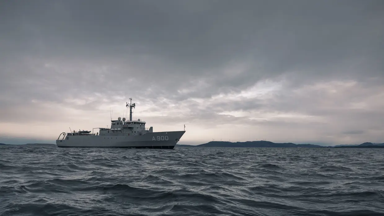 Een grijs schip vaart op zee met een kustlijn in de verte 