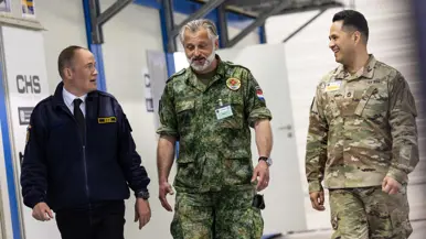 Links: Een man in pak met bril en identiteitskaart werkt op een laptop. Een vrouw in militair uniform met een medisch embleem staat naast hem. Rechts: drie mannen lopen glimlachend door een gang: een in donkerblauw uniform, een in Nederlands militair tenue met medisch embleem, en een in Amerikaans legeruniform. 