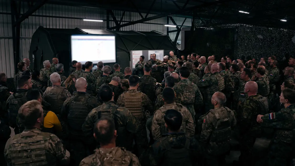 Tientallen militairen op de rug gezien drommen samen rond een tafel met daarop een kaart. Er hangt ook een groot projectiescherm met de laatste info.