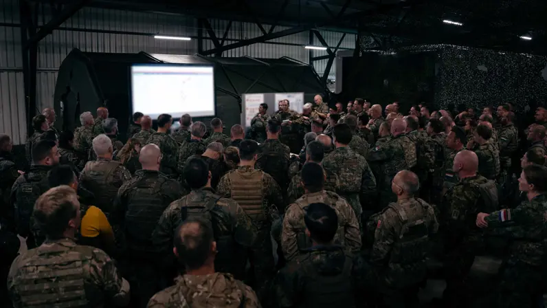 Tientallen militairen op de rug gezien drommen samen rond een tafel met daarop een kaart. Er hangt ook een groot projectiescherm met de laatste info.