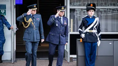 Op de ene foto de twee generaals die naast elkaar lopen. Beiden brengen ze de militaire groet. Daarnaast het duo, staand in de houding, op een kleine verhoging. Ook hier brengen ze de militaire groet. Een groep opgestelde marechaussees groet terug, te zien vanaf de rug. Op de achtergrond is een grijs gebouw zichtbaar.