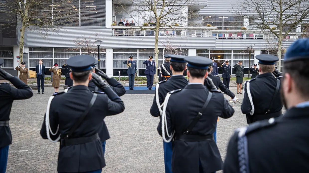 Op de ene foto de twee generaals die naast elkaar lopen. Beiden brengen ze de militaire groet. Daarnaast het duo, staand in de houding, op een kleine verhoging. Ook hier brengen ze de militaire groet. Een groep opgestelde marechaussees groet terug, te zien vanaf de rug. Op de achtergrond is een grijs gebouw zichtbaar.
