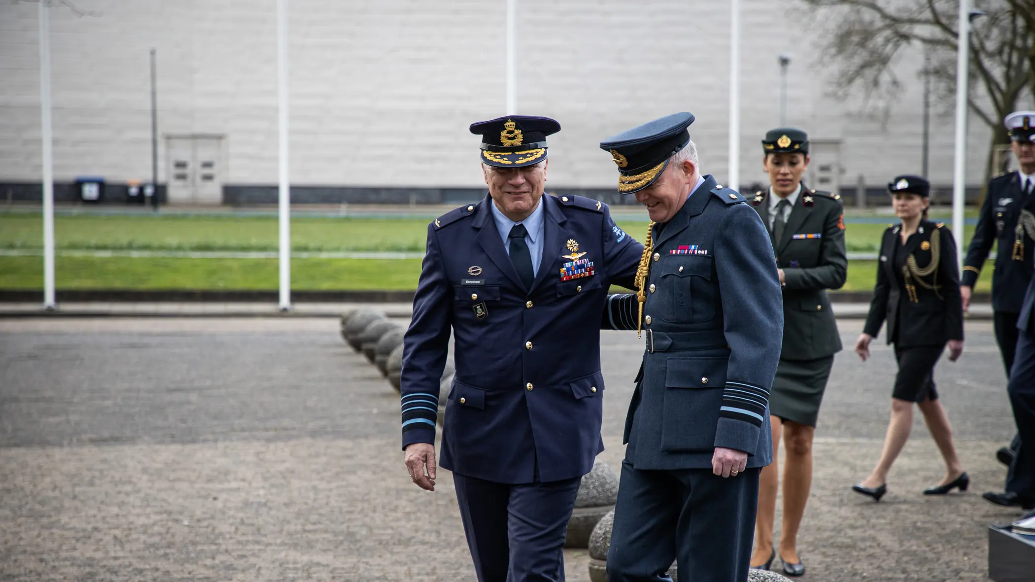 Generaal Onno Eichelsheim loopt lachend naast zijn Britse collega. De twee, in donkerblauw militair uniform, hebben een hand op elkaars rug.