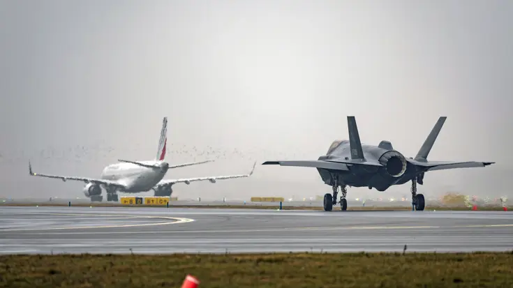 Een taxiënde F-35 rijdt over de landingsbaan op Schiphol. In de verte is een passagiersvliegtuig te zien. De lucht is grijs.
