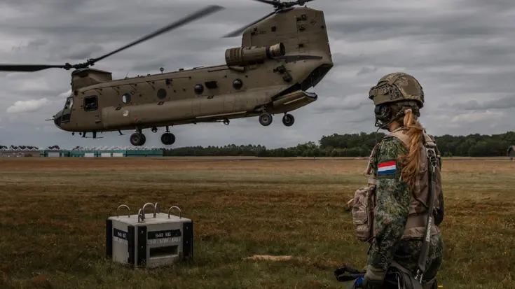 Prinses Amalia, gehuld in gevechtstenue, kijkt naar een Chinook-transporthelikopter de niet ver bij haar vandaan in de lucht hangt.