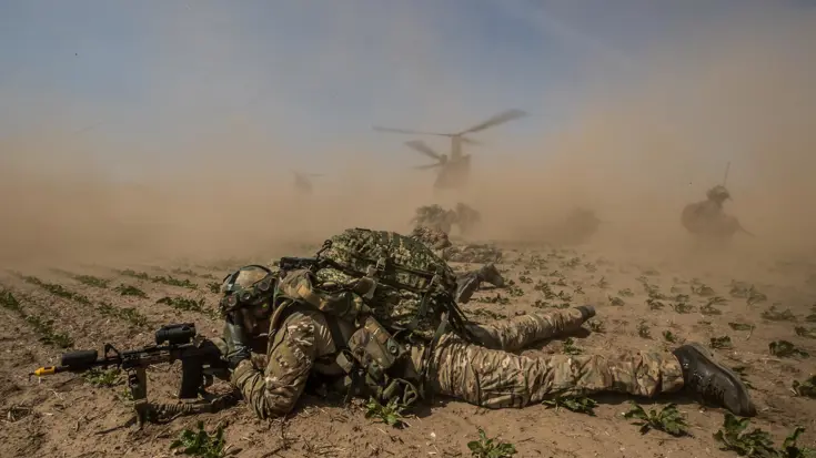 Soldaat ligt in veld met geweer voor zich. Op de achtergrond staat een helikopter met draaiende motor. Zand en stof waait alle kanten op.