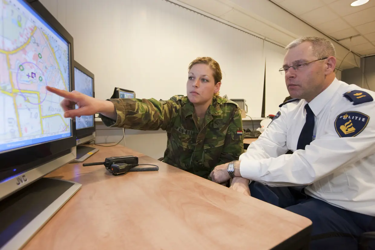 Een vrouwelijke militair en een politieman kijken naar een beeldscherm waar een plattegrond op te zien is.