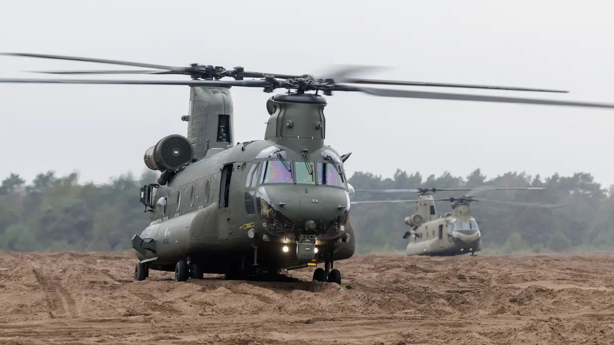 Twee donkergroene militaire helikopters op een zandig veld. De voorste draait met draaiende rotor naar de kijker; de tweede staat erachter. Achteraan bomen onder een bewolkte lucht.