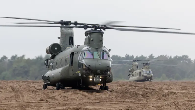 Twee donkergroene militaire helikopters op een zandig veld. De voorste draait met draaiende rotor naar de kijker; de tweede staat erachter. Achteraan bomen onder een bewolkte lucht.