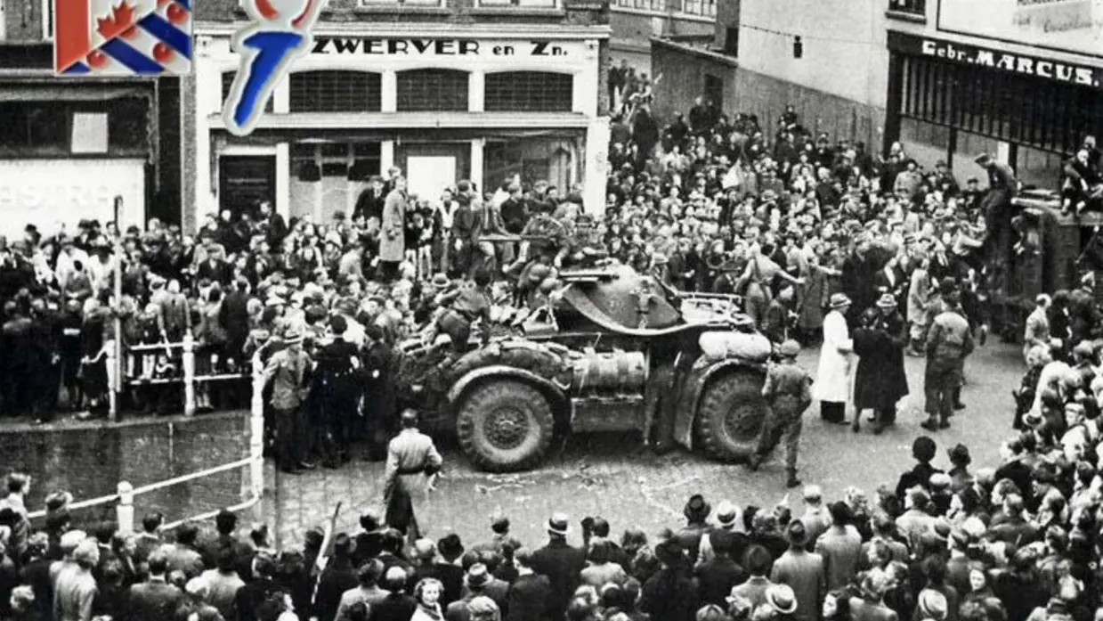 Een grote menigte omringt een militaire pantserwagen op een straat, met gebouwen en een bord met de Canadese vlag op de achtergrond. De foto lijkt historisch.