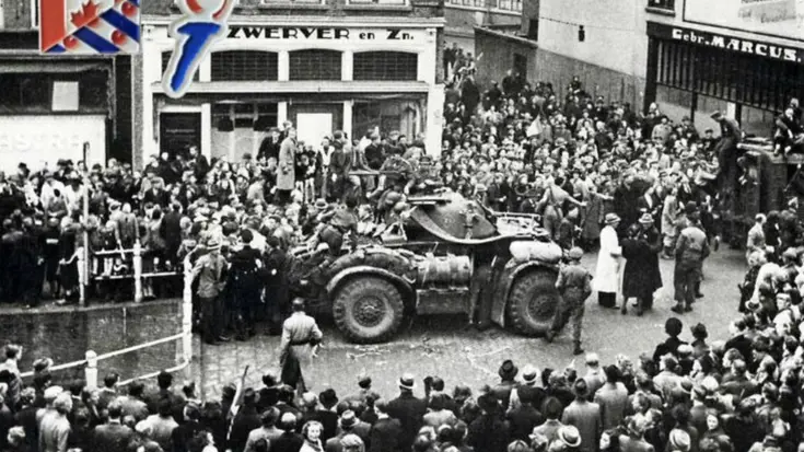 Een grote menigte omringt een militaire pantserwagen op een straat, met gebouwen en een bord met de Canadese vlag op de achtergrond. De foto lijkt historisch.