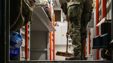 Een persoon in camouflagebroek en laarzen staat op een metalen platform en veegt met een bezem. Industriële omgeving met oranje frames en apparatuur.