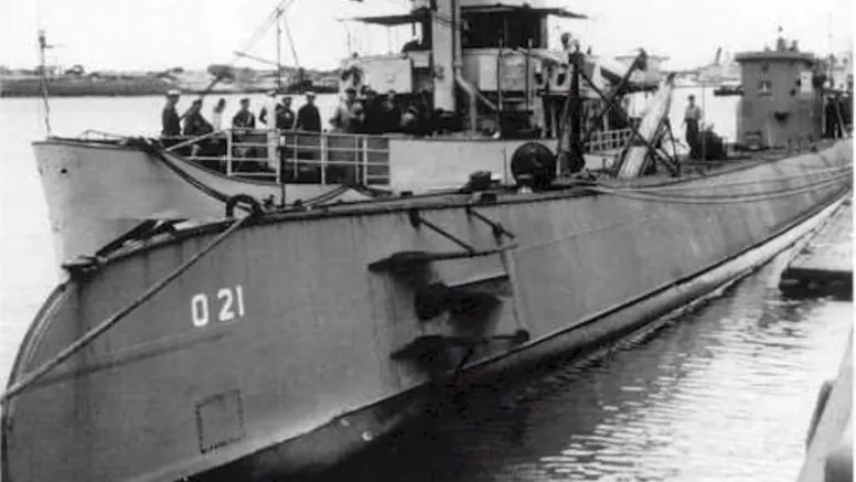 Onderzeeboot met nummer 021 op de romp, afgemeerd in een haven. Bemanning op het dek, met kraan en geschutskoepel. Industriële achtergrond, met andere schepen in kalm water.