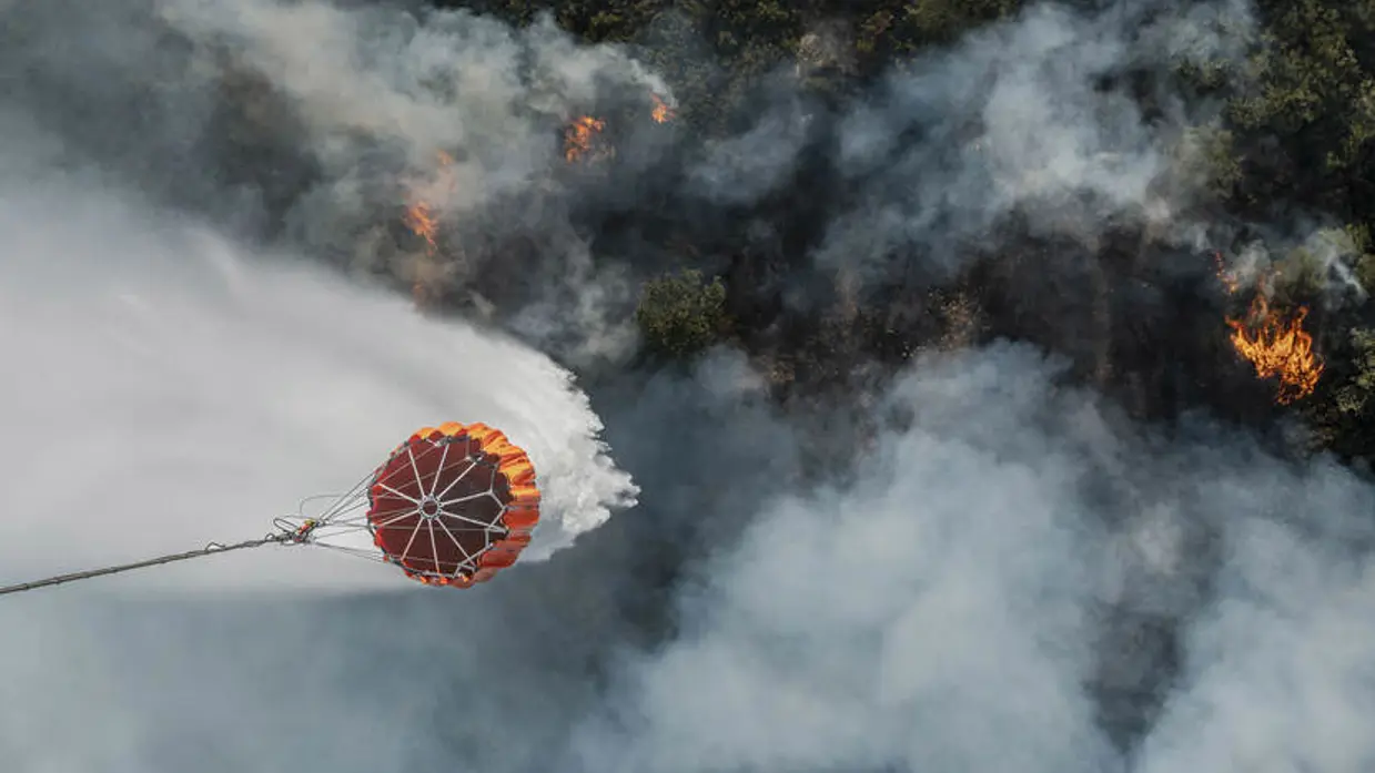 Foto vanuit Chinook-helikopter recht naar beneden. Een bambi bucket heeft zojuist water gedropt op brandend terrein.