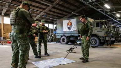 Vier soldaten in groene camouflageuniformen staan in een garage; één wijst met een stok naar een kaart op de vloer, anderen bekijken papier. Militaire voertuigen op de achtergrond.