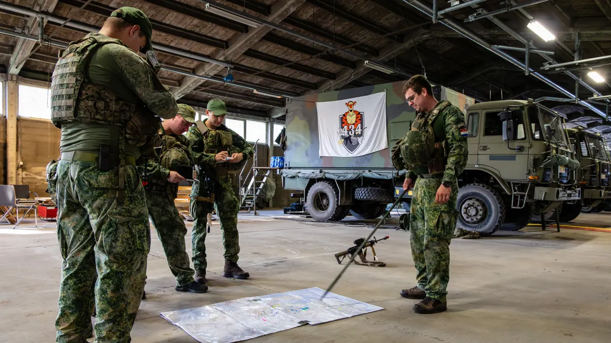 Vier soldaten in groene camouflageuniformen staan in een garage; één wijst met een stok naar een kaart op de vloer, anderen bekijken papier. Militaire voertuigen op de achtergrond.