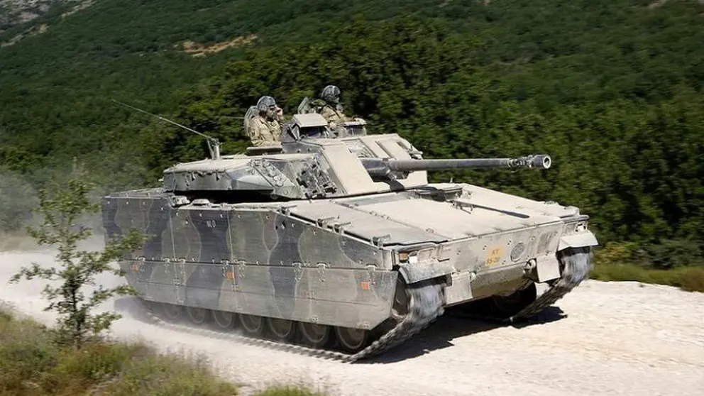 Een CV90 gevechtvoertuig rijdt op een zandpad. Op de achtergrond een groen begroeide heuvelrug.