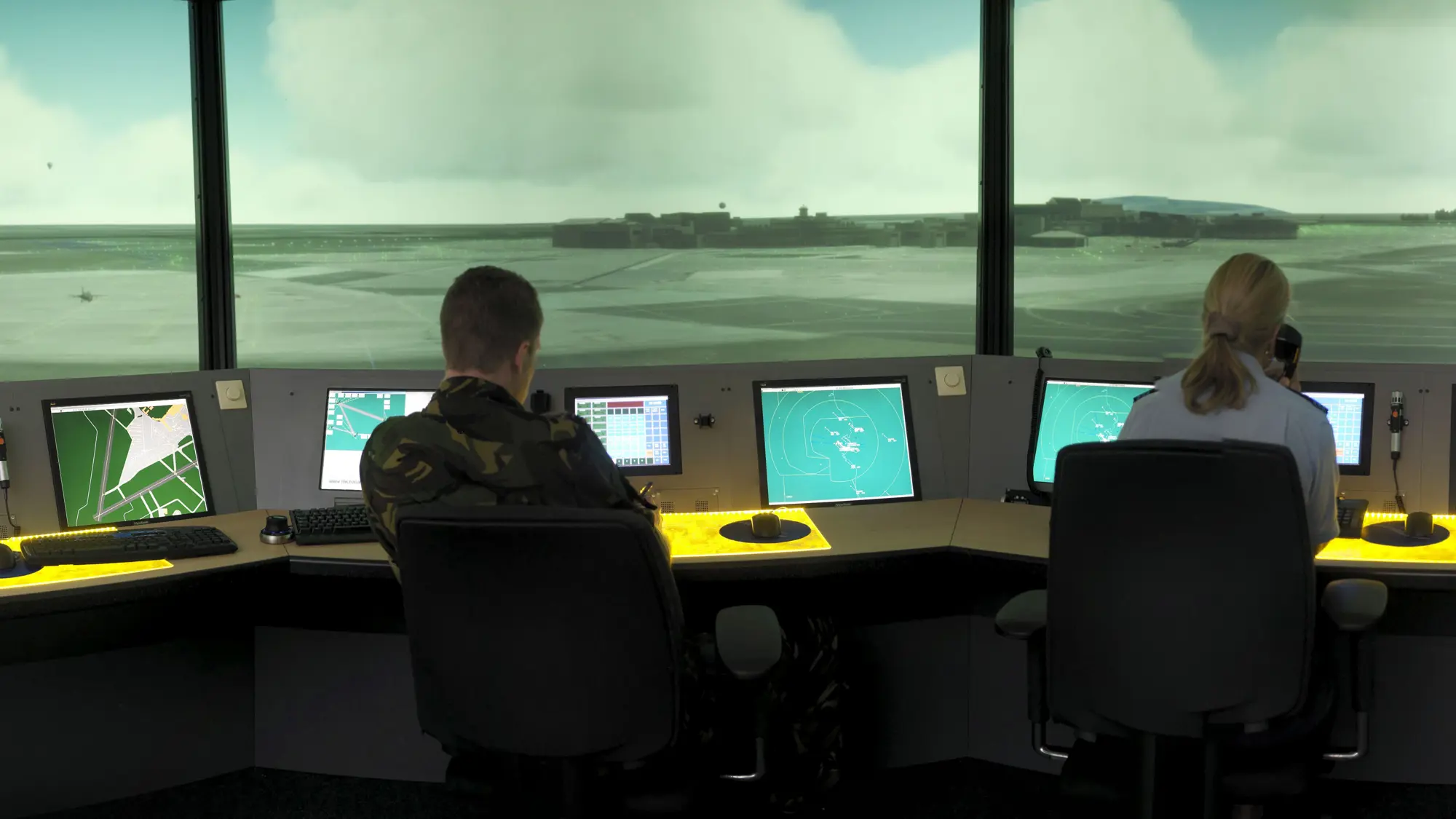 Twee militairen van de luchtverkeersleiding kijken uit op de start- en landingsbanen vanachter hun computerschermen.