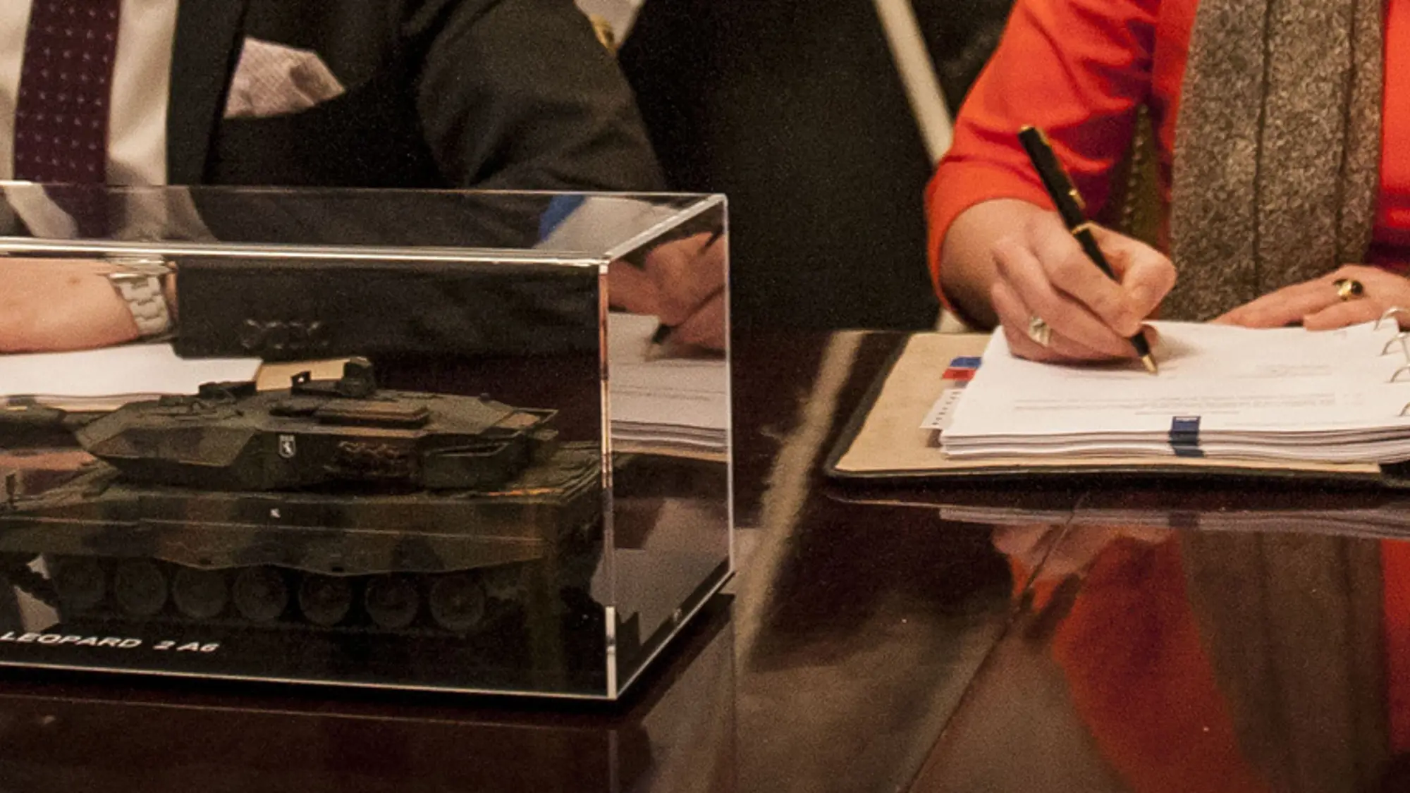Close-up van de handen van twee personen die elk een document ondertekenen. Op de voorgrond staat een schaalmodel van een tank in een plexiglas doosje op tafel.