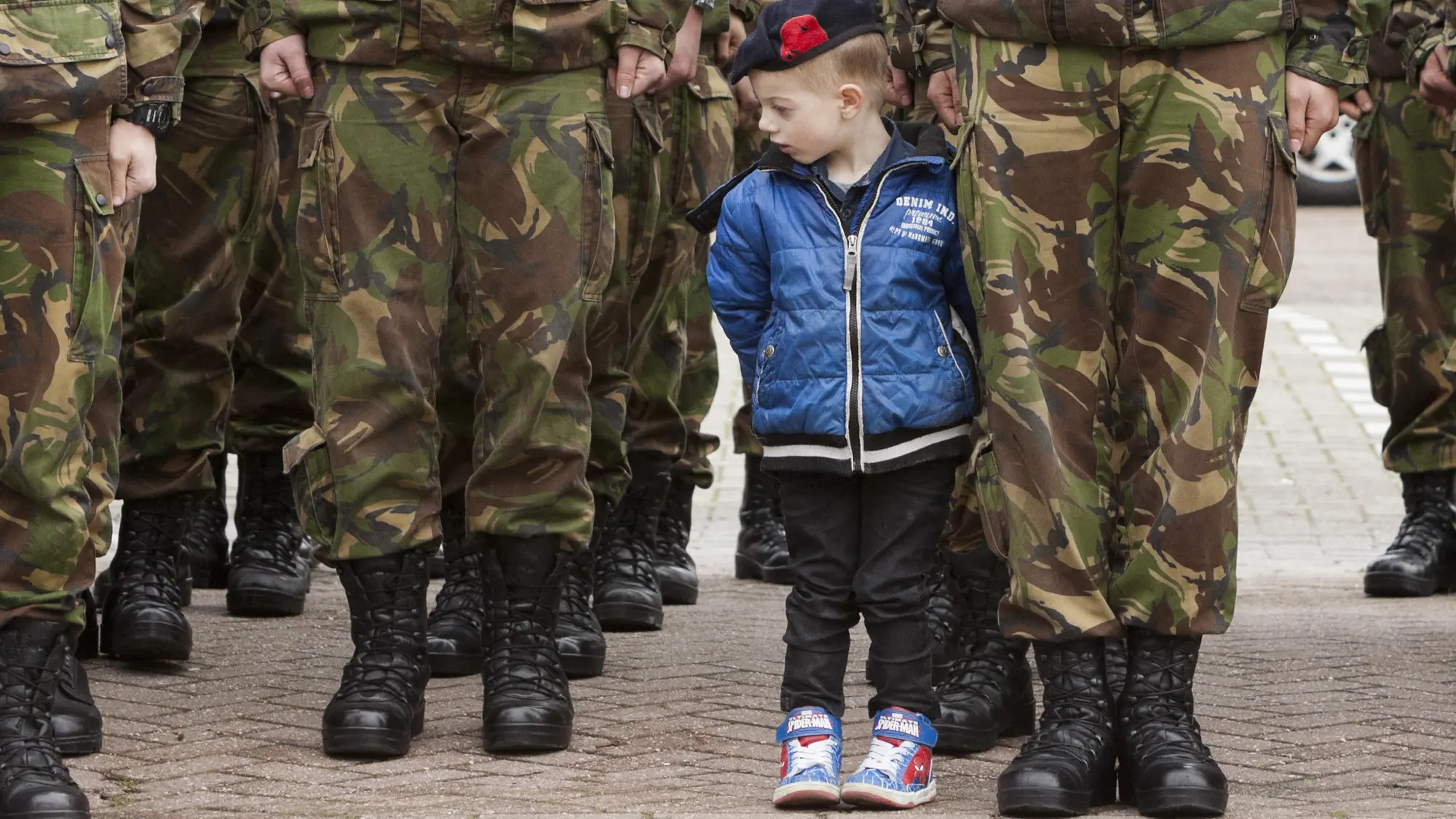 Klein jongetje met kinderbaret op staat tussen benen van militairen in uniform en met legerkisten aan.