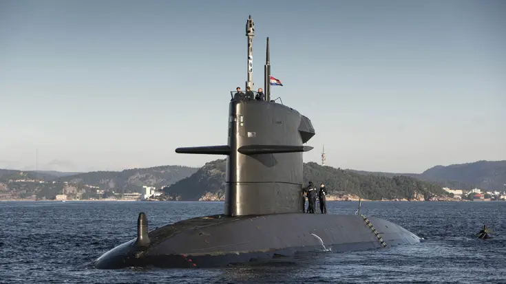 Een onderzeeboot komt boven water en hijst de Nederlandse vlag. Bovenop het vaartuig zijn militairen van de marine met elkaar in gesprek. Op de achtergrond een bergachtige kustlijn.