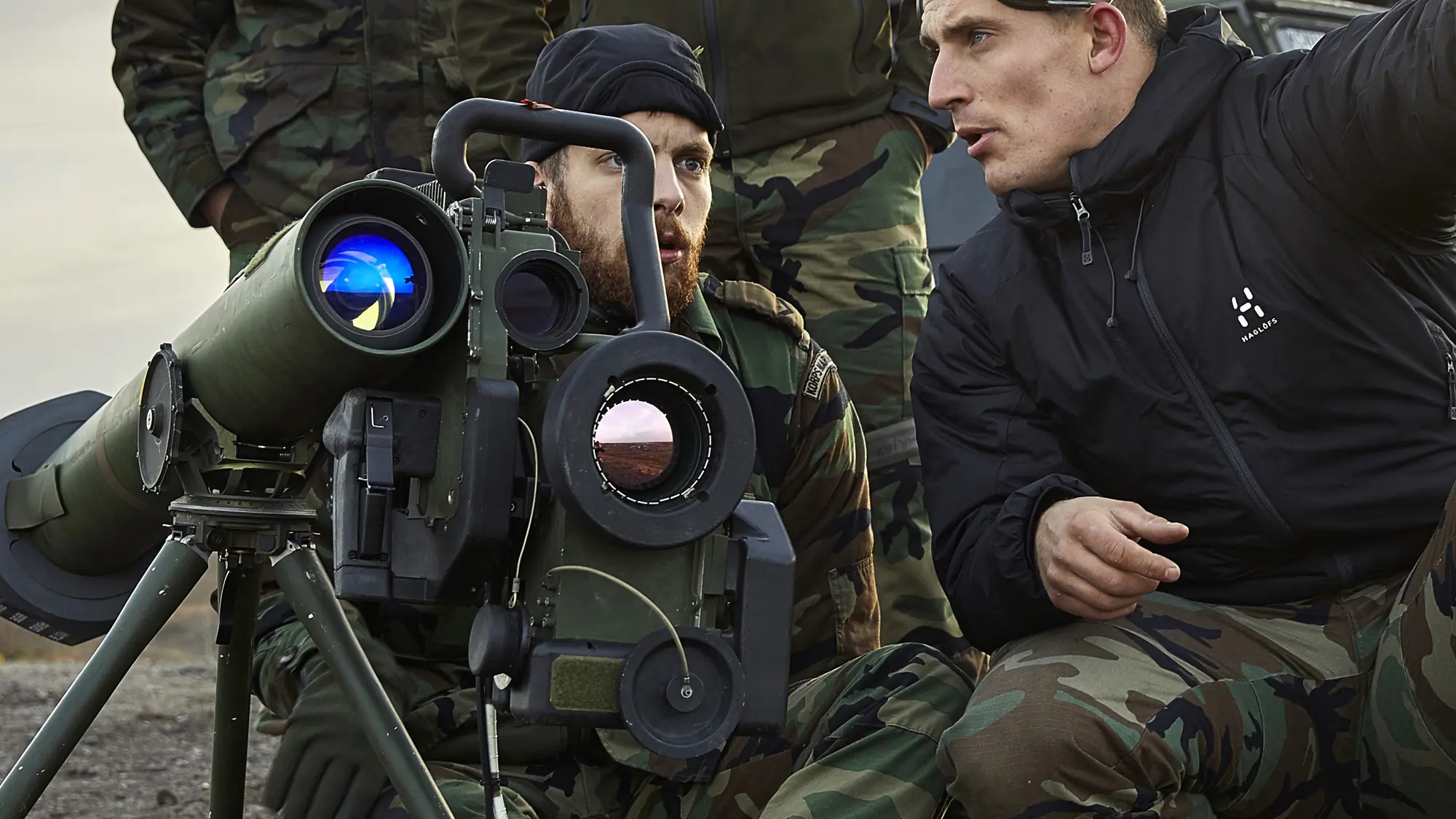 Twee militairen zitten gehurkt naast militair materieel tijdens uitleg en overleg.