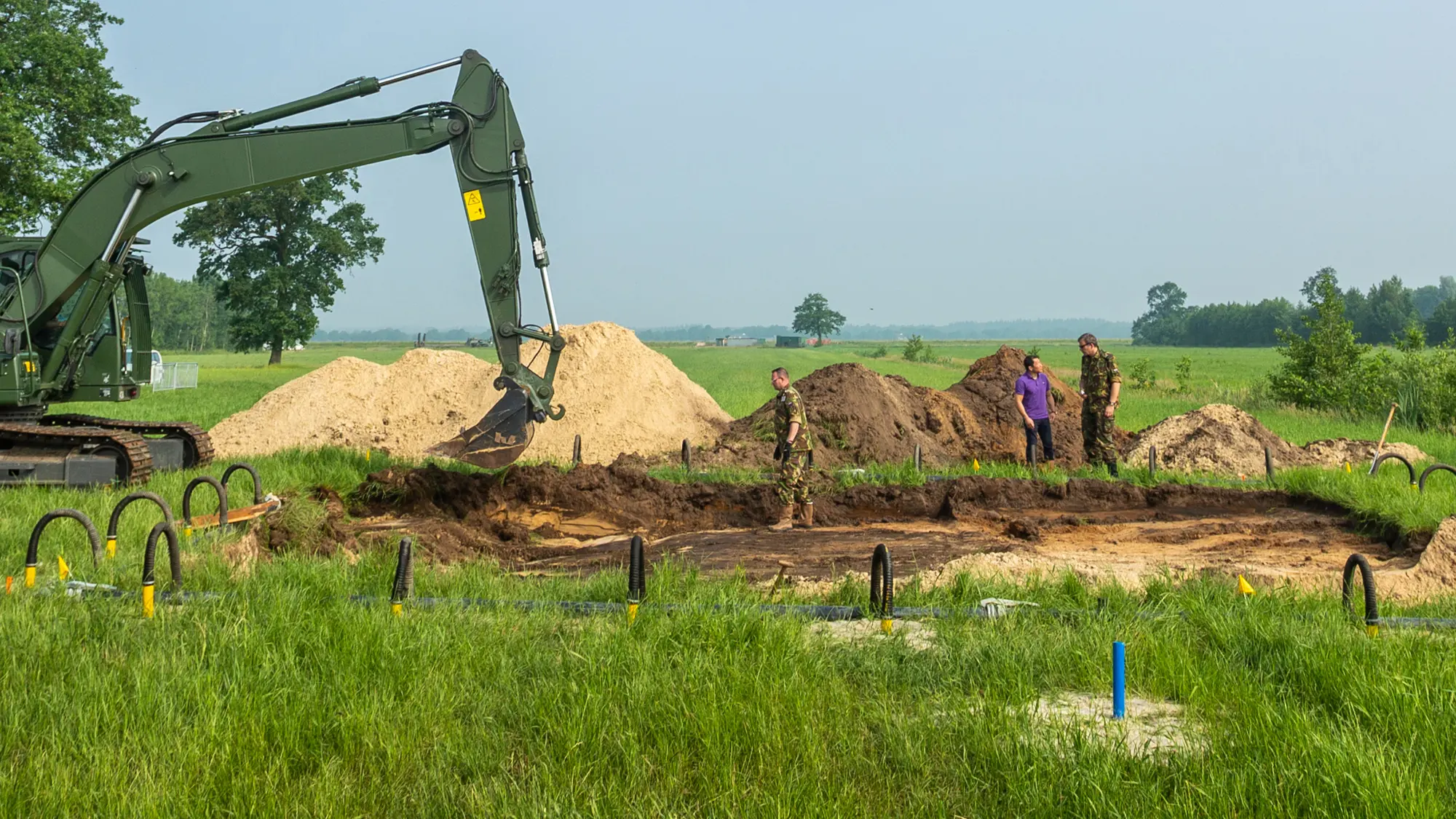 Graafmachine graaft in grasveld tijdens berging en identificatie oorlogsslachtoffers.