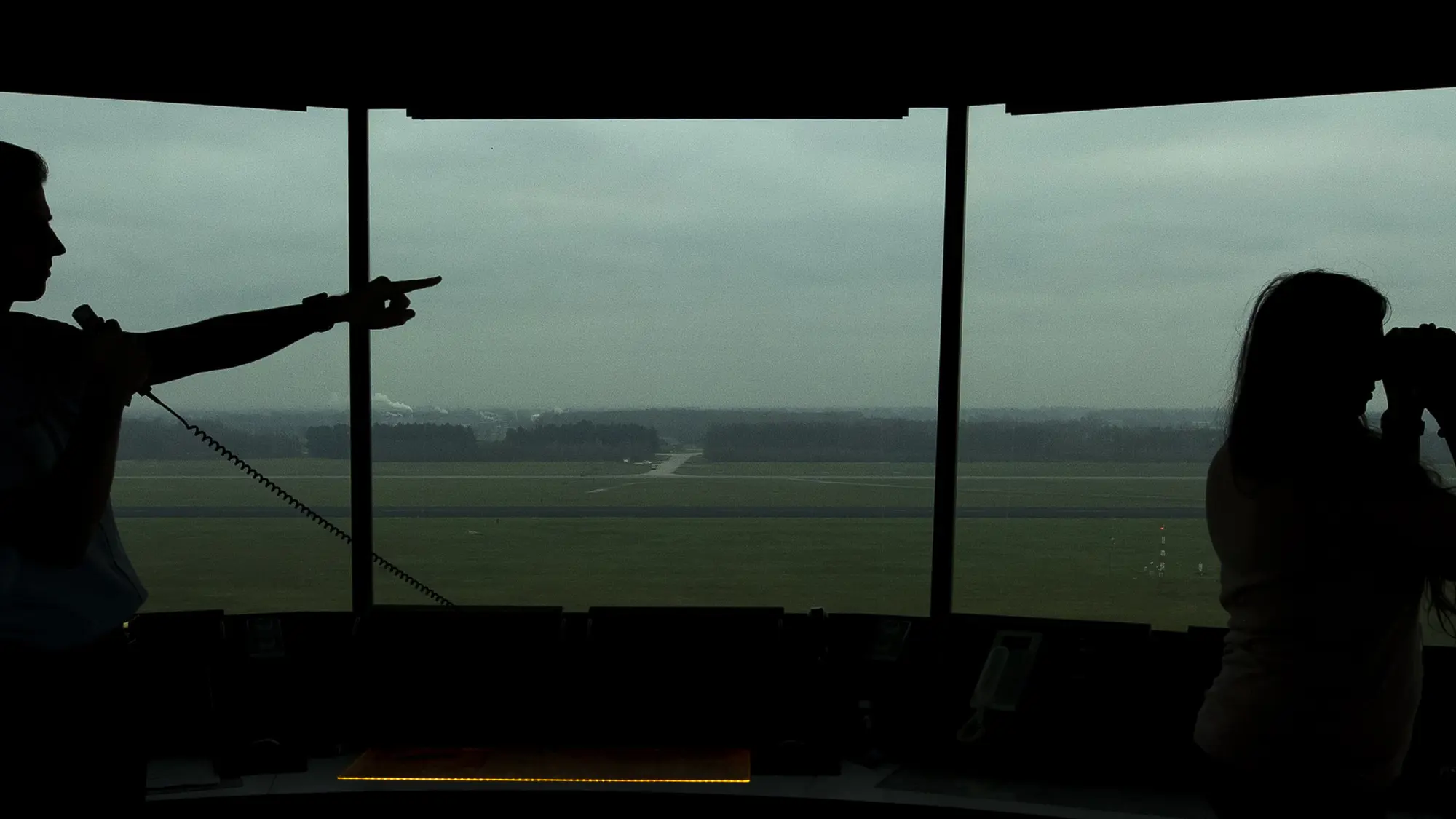 Silhouet van twee medewerkers in luchtverkeerstoren waarvan één met een verrekijker kijkt terwijl de ander iets aanwijst.