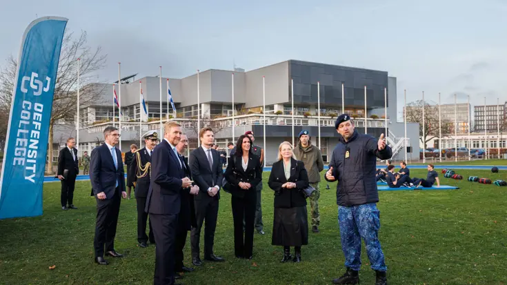De Koning en minister staan met een aantal andere mensen bij de vlag van Defensity College.