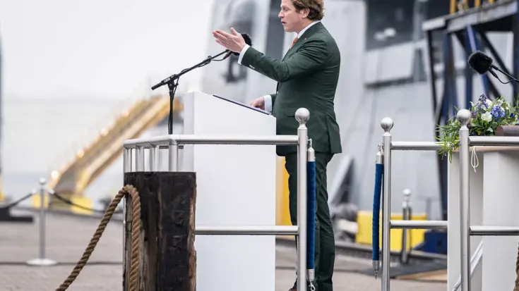 Staatssecretaris Gijs Tuinman in groen pak achter katheder op kade. Marineschip is vaag op de achtergrond zichtbaar.