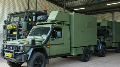 Drie groene legervoertuigen staan onder een dak op een betonnen vloer.